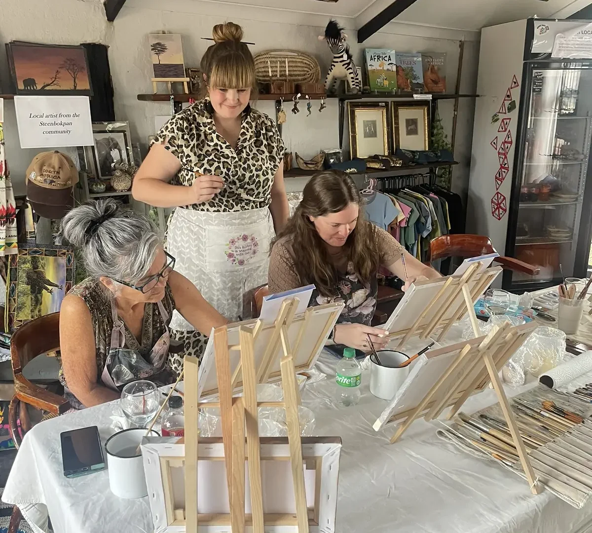 A tutor guiding participants who are painting around a busy workshop table.