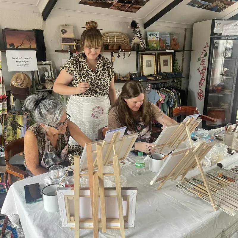 A tutor guiding participants who are painting around a busy workshop table.