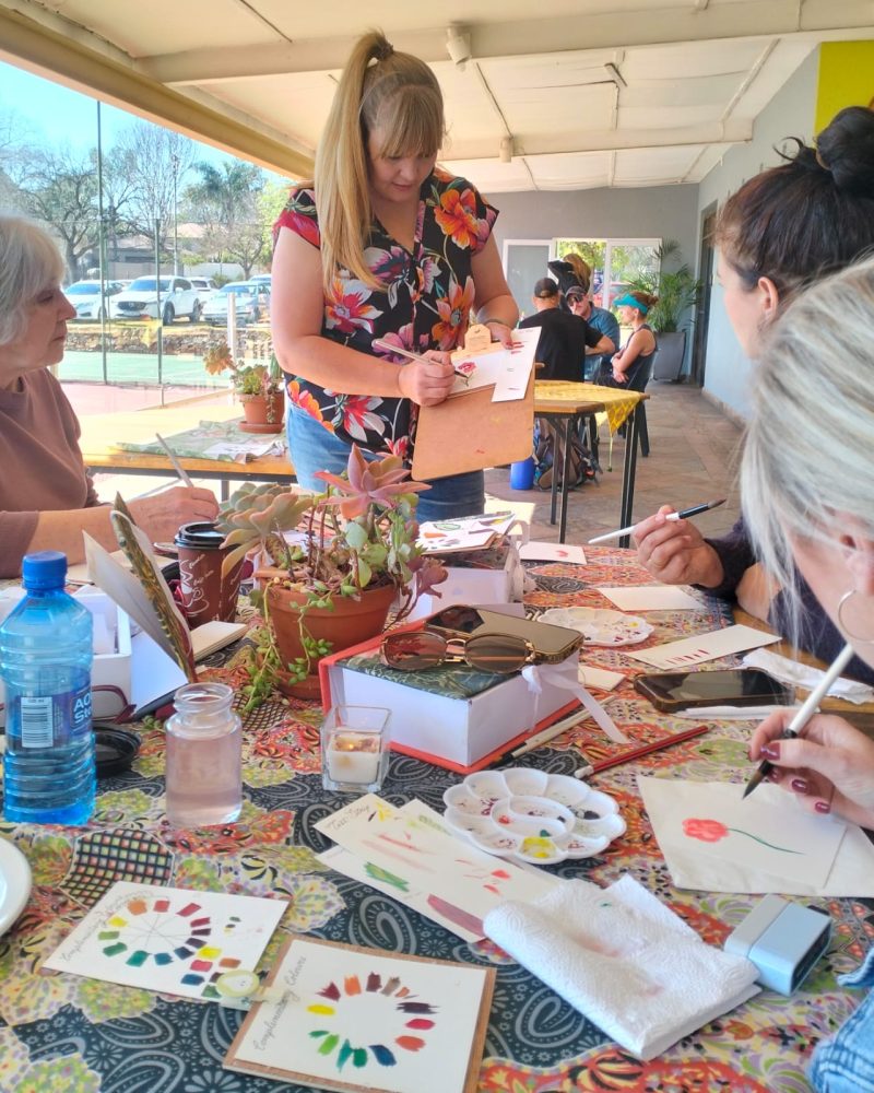 A tutor guiding participants who are painting around a busy workshop table.