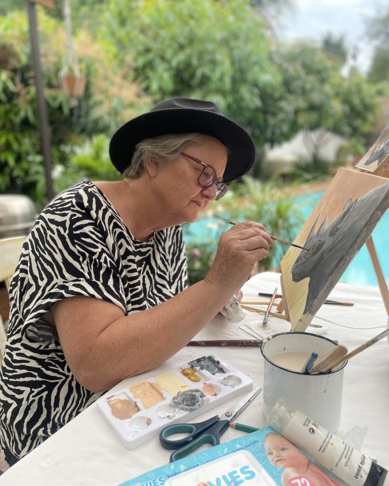 A participant in a hat painting on a tabletop easel in a garden setting.