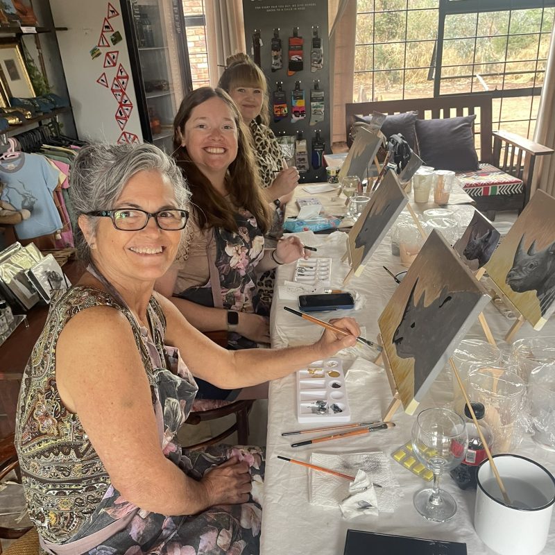 Smiling participants painting at tabletop easels along a long table.