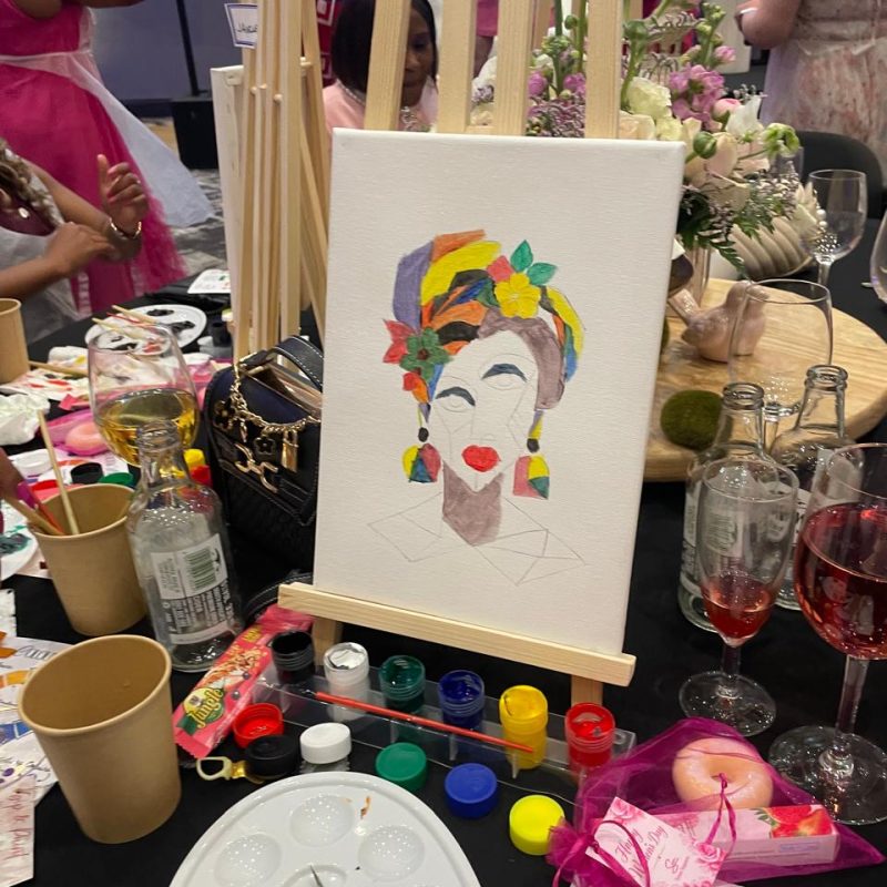 A colourful portrait painting on a tabletop easel among paints and wine glasses at a paint-and-sip table.